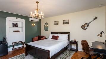 A bedroom with a bed, a desk, a chair, a guitar, a chandelier, and a small shelf with pictures.