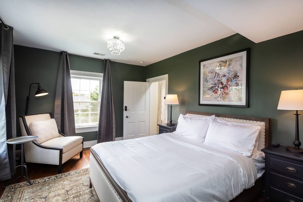 A bedroom with a large bed, a chair, a small table, a nightstand, and a chandelier.