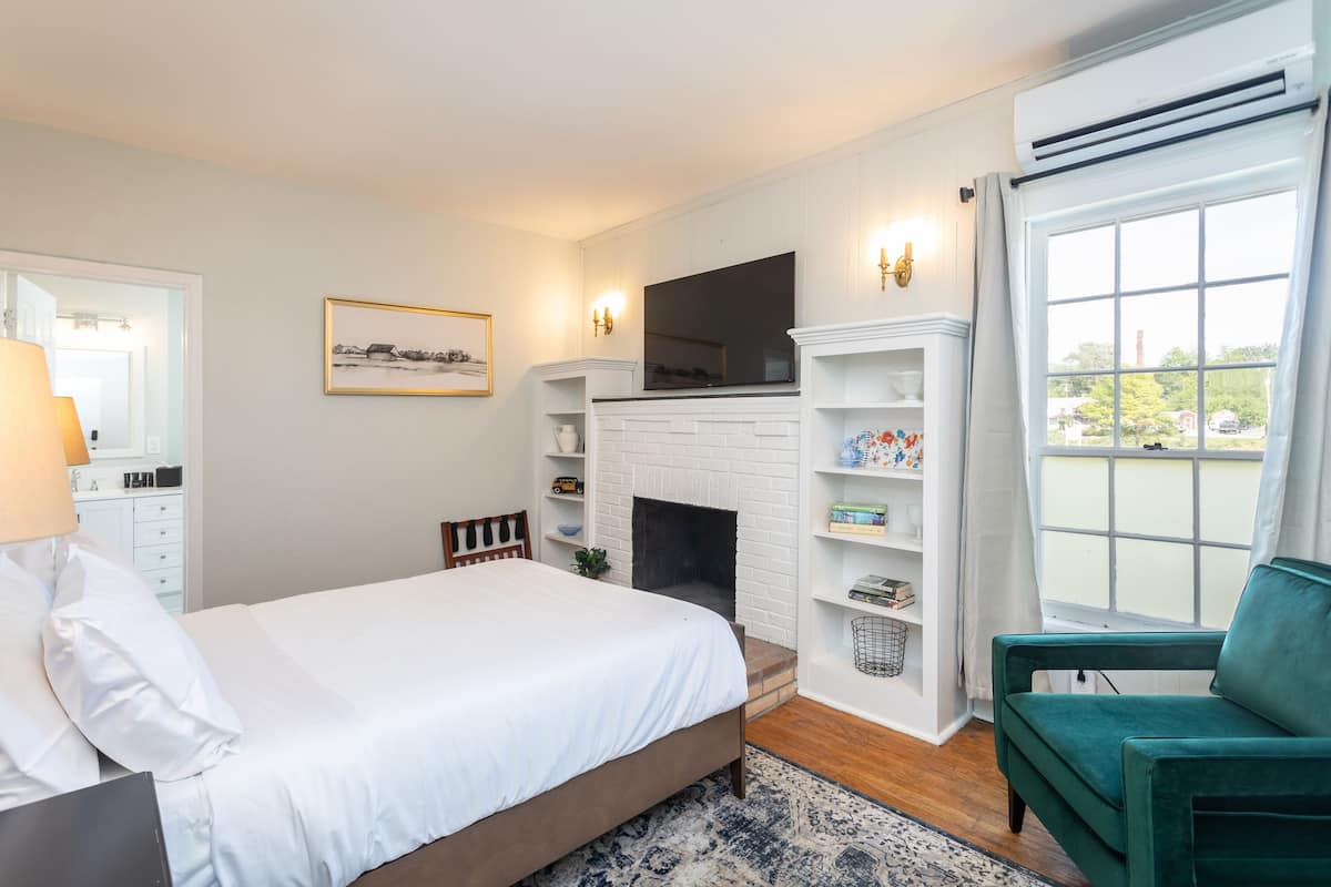A bedroom with a bed, a fireplace, a TV mounted above it, a green armchair, and a large window with a view.