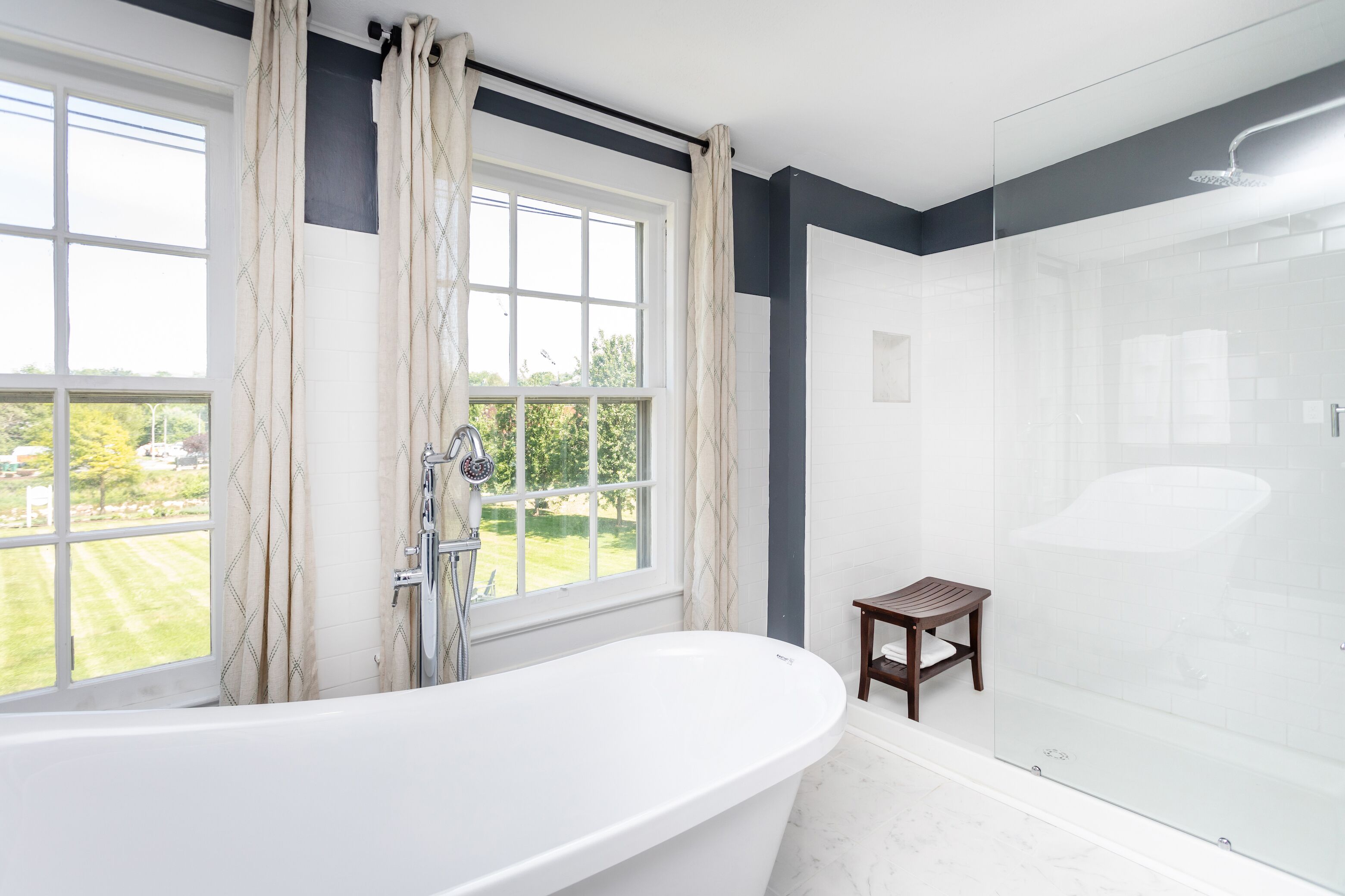 A modern bathroom with a freestanding bathtub, a glass-enclosed shower, and a view of a green landscape through large windows.