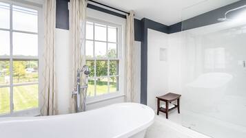 Un baño moderno con una bañera independiente, ducha con mampara de vidrio y vistas a un paisaje verde a través de amplios ventanales.