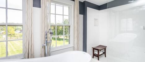 A modern bathroom with a freestanding bathtub, a glass-enclosed shower, and a view of a green landscape through large windows.