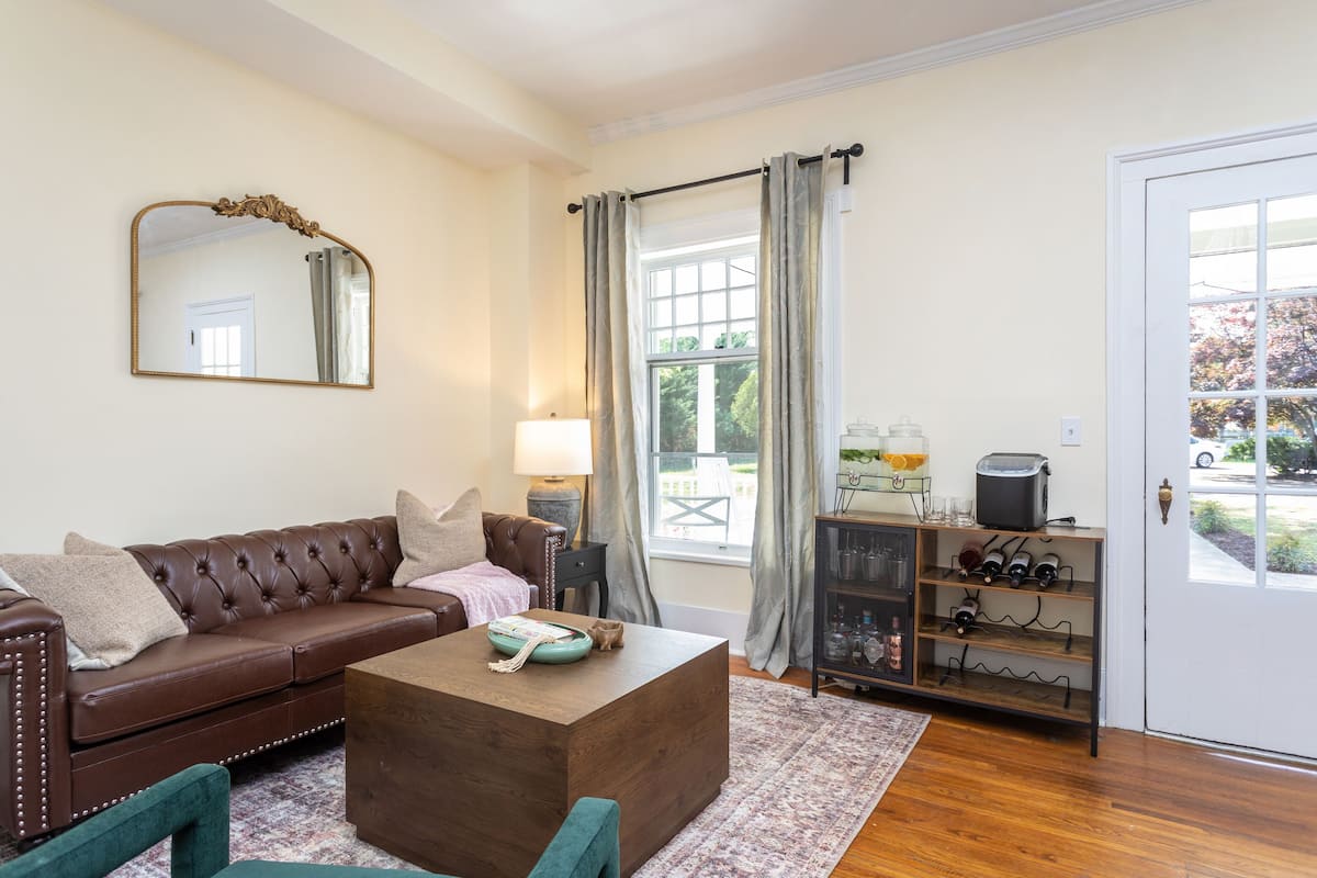 A living room with a brown leather sofa, a wooden coffee table, a mirror, a lamp, a wine rack, and a view of the outdoors through a window.