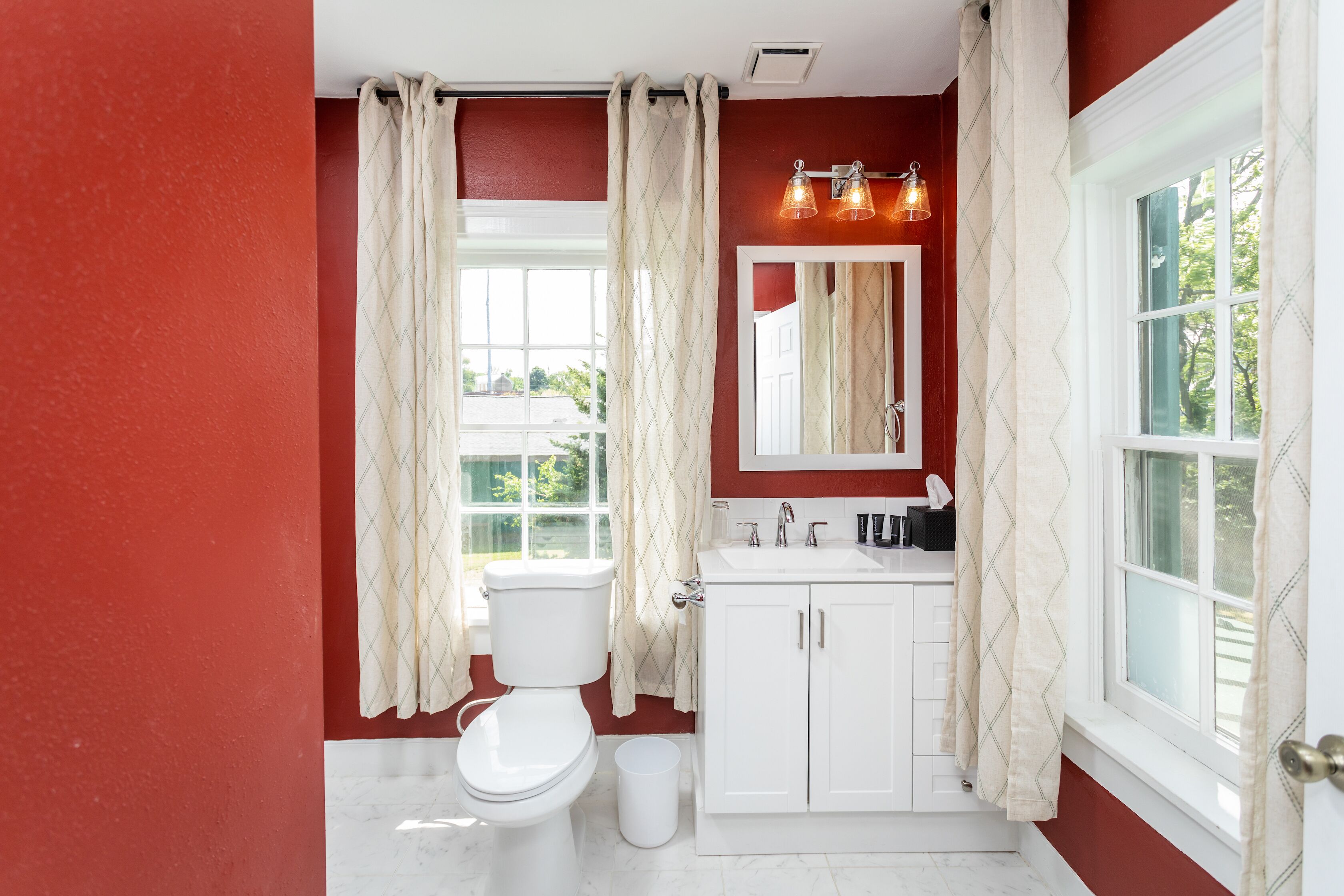 A bathroom with a white toilet, white cabinets, a white sink, and a window with curtains.