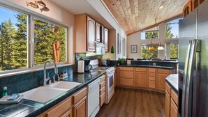 A modern kitchen with wooden cabinets, a stainless steel refrigerator, and a large window offering a view of trees. - Whispering Pines in Tahoe Vista (Tahoe Vista)
