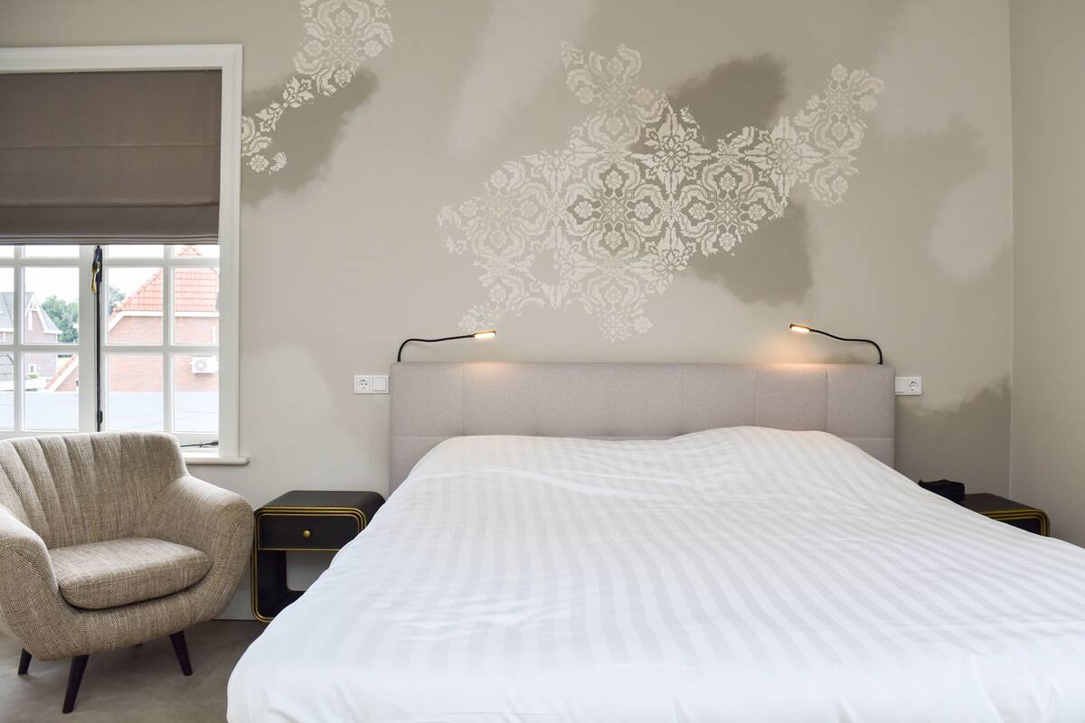 A bedroom with a large bed, a chair, a nightstand, and a wall with a decorative pattern.