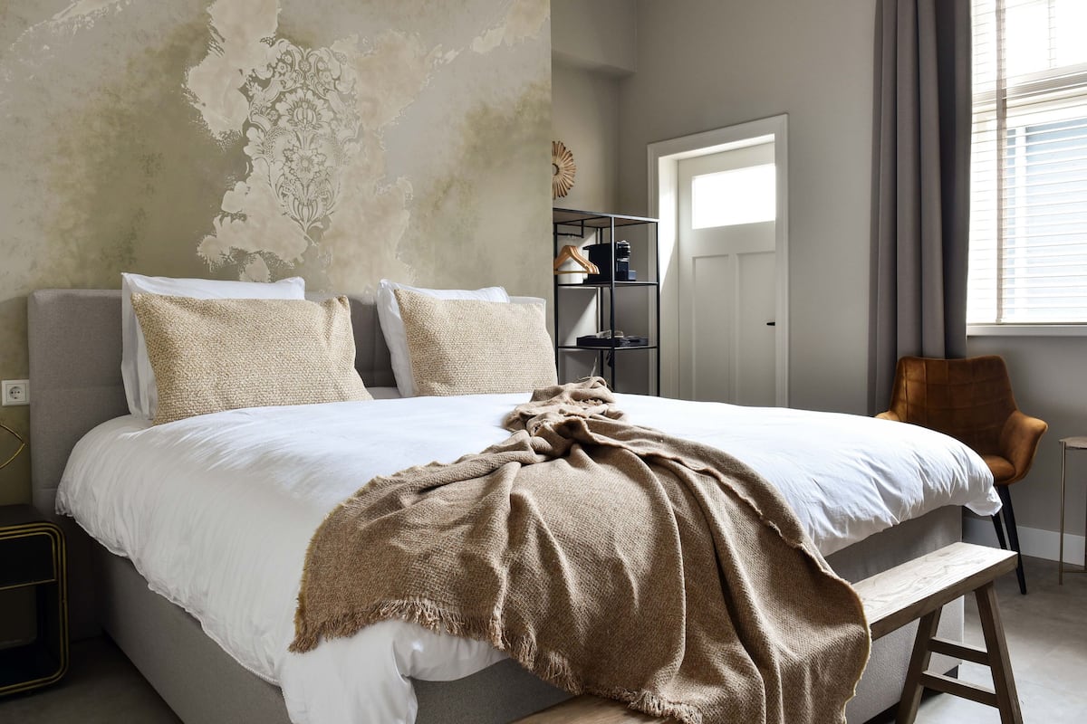A bedroom with a large bed, a bench, a chair, a wardrobe, and a door.