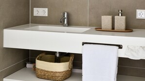 Une salle de bain avec un miroir rond, un lavabo blanc, deux distributeurs de savon, un porte-serviettes et un panier en osier placé sous le lavabo.