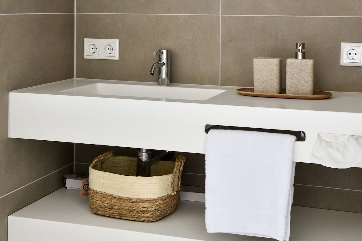 A bathroom with a round mirror, a white sink, two soap dispensers, a towel rack, and a woven basket under the sink.