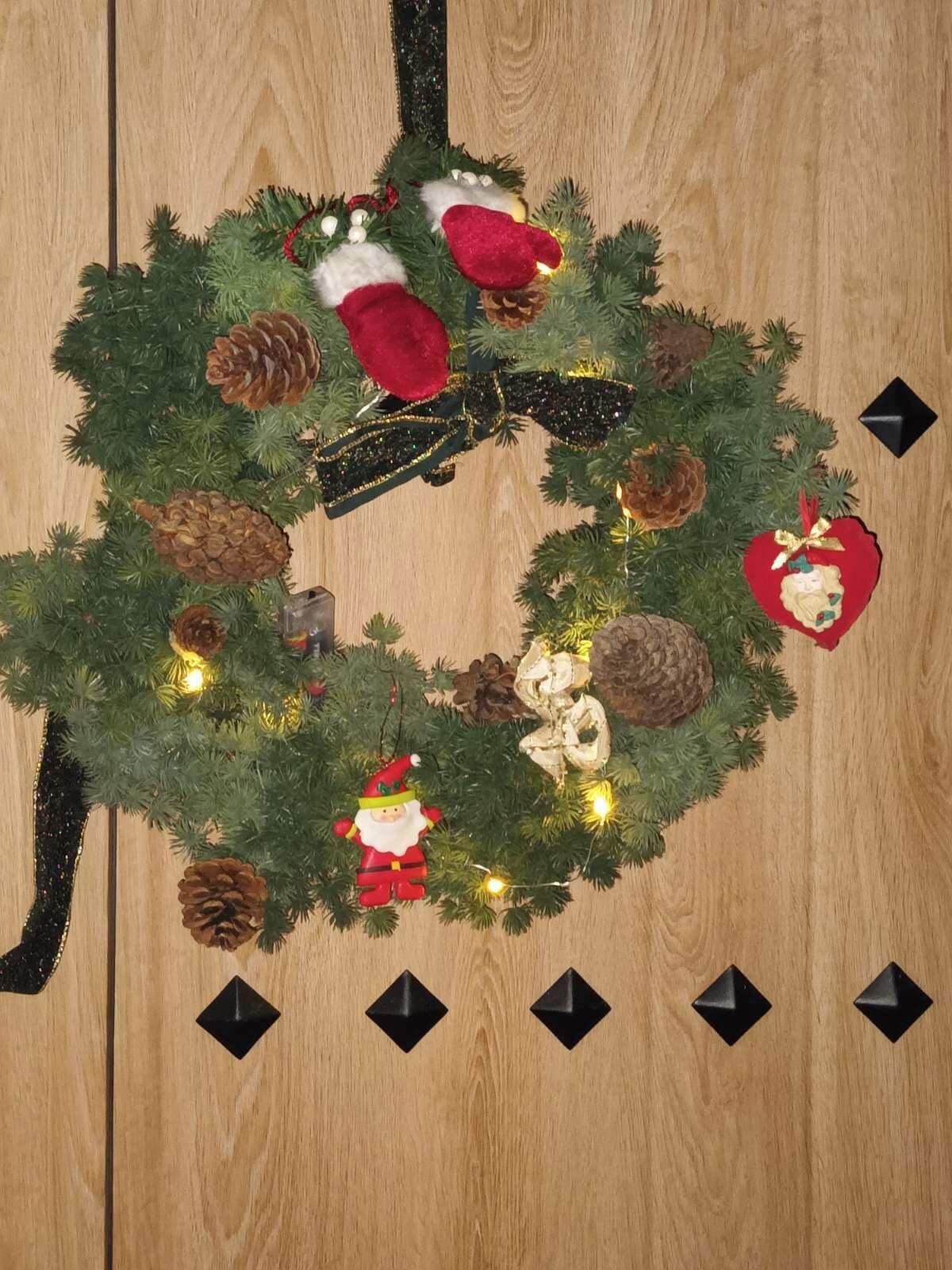 Un adorno navideño con un lazo de piñas, un adorno de Papá Noel y luces sobre una superficie de madera.