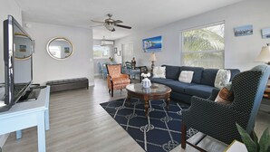Een moderne woonkamer met een blauwe bank, een gestreept fauteuil en een salontafel. Er is een televisie op een dressoir, een ronde spiegel en een plafondventilator.
