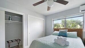 Een slaapkamer met een bed, een plafondventilator, een raam met uitzicht op palmbomen en een kledingkast met planken.