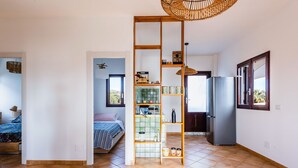 A spacious room with a kitchen area, a bed with a blue and white striped bedspread, and a wooden shelf unit with kitchen items. - Villetta Sirena a Fontane Bianche (Syracuse)