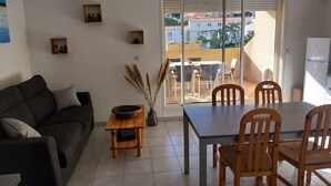 A living room with a sofa, a dining table with chairs, a small wooden table with a bowl, and a vase with dried plants. There is a sliding glass door leading to an outdoor balcony.