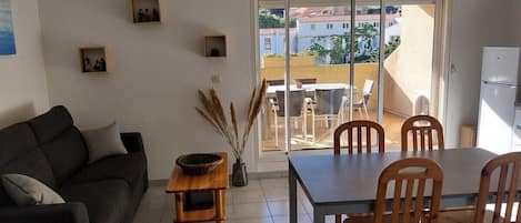Uma sala de estar com sofá, mesa de jantar com cadeiras, uma pequena mesa de madeira com uma tigela e um vaso com plantas secas. Há uma porta de vidro deslizante que dá para uma varanda externa.