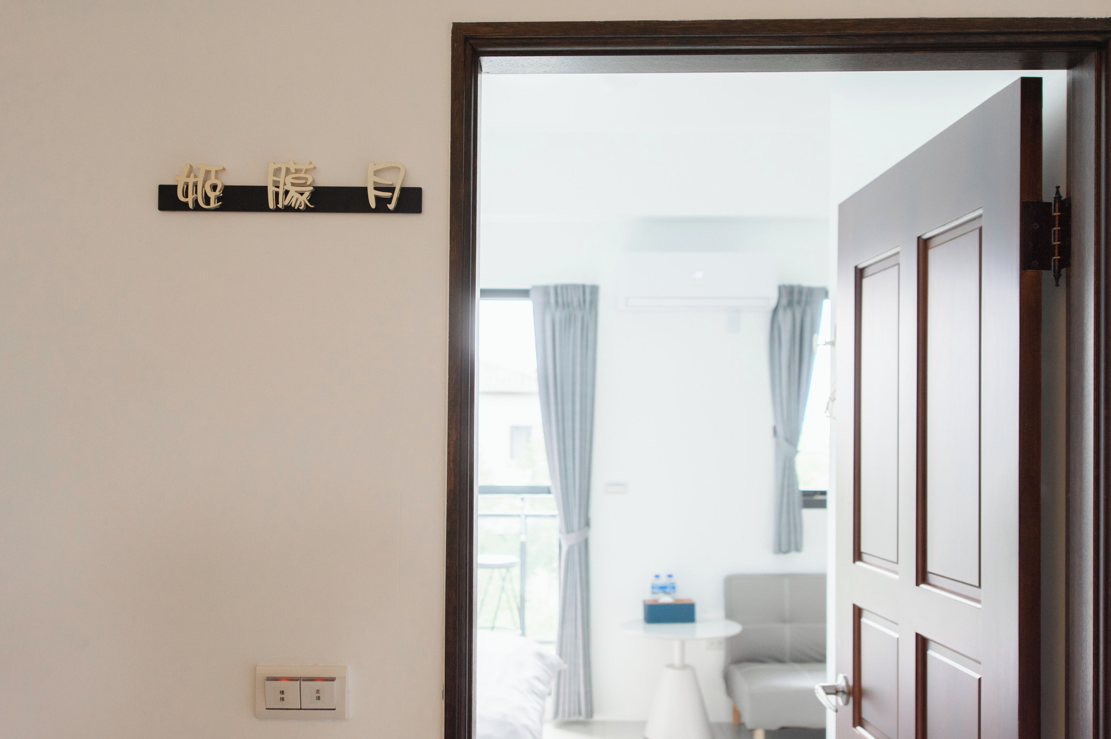 A hotel room entrance with a wooden door, a white wall, and a small table with a chair.