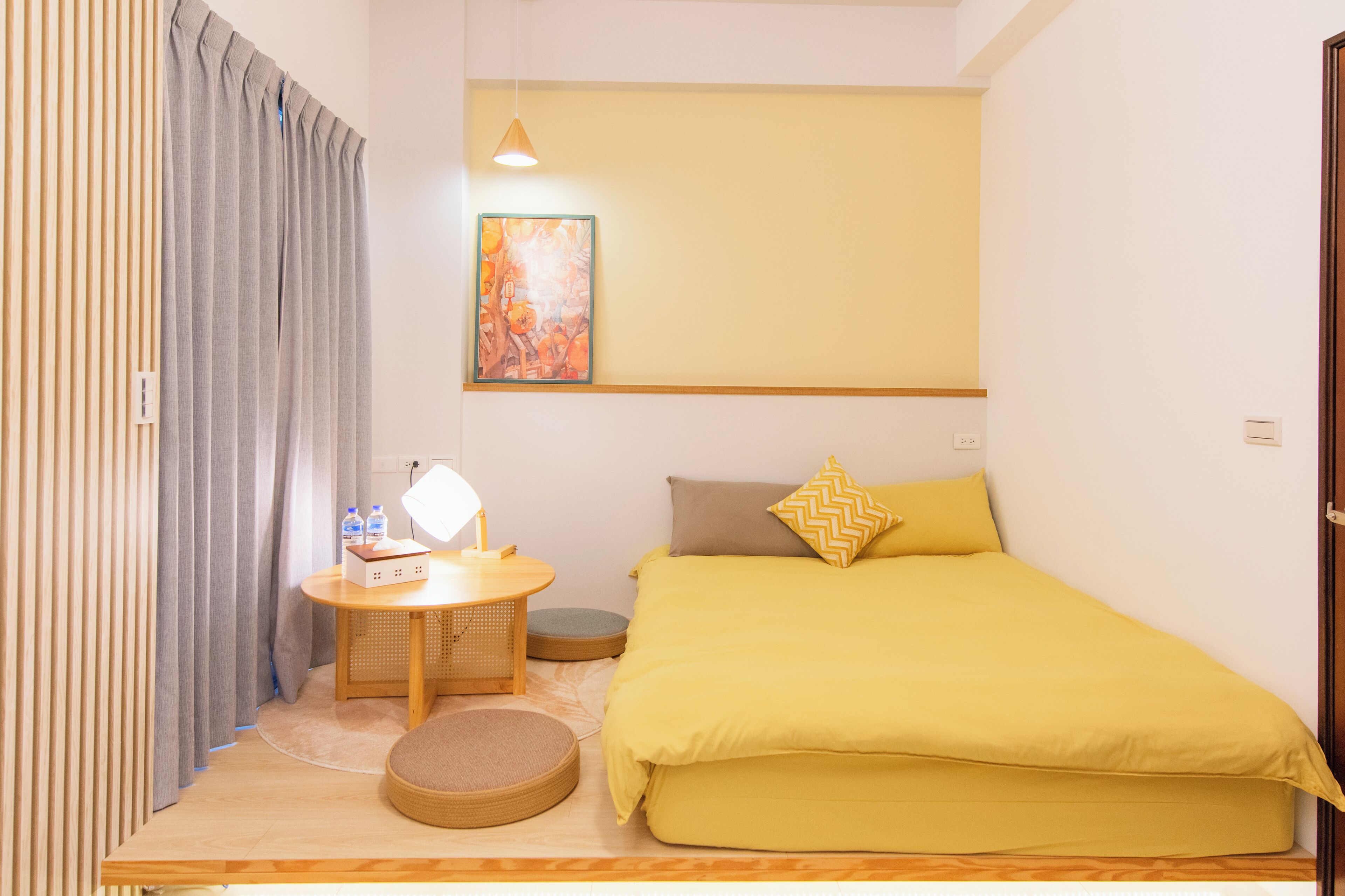 A compact room with a bed, a small table with a lamp, a round cushion, and a wall-mounted painting.