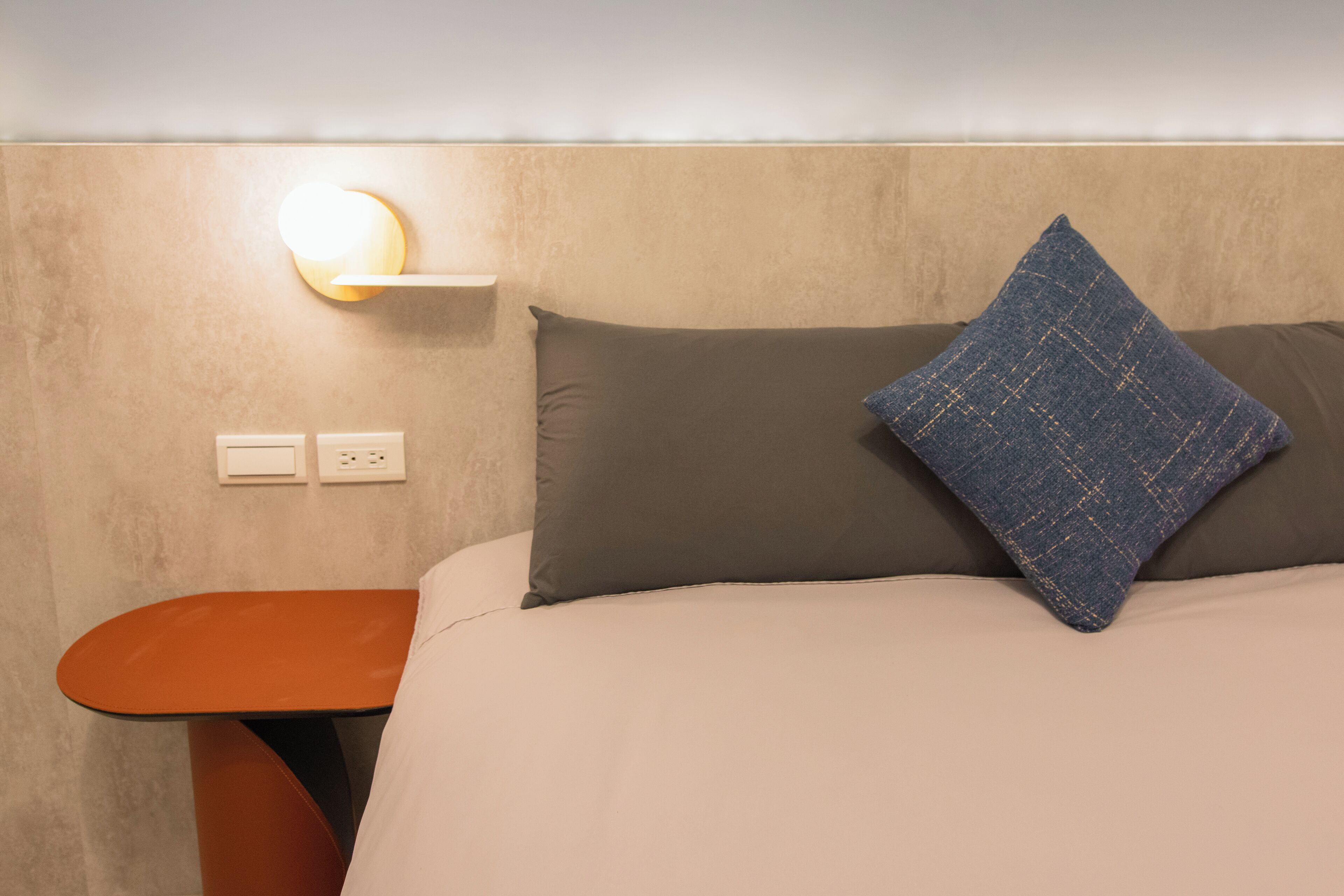 A bed with a beige mattress, a dark gray pillow, and a blue textured pillow. There is a small orange table beside the bed with a light on it.