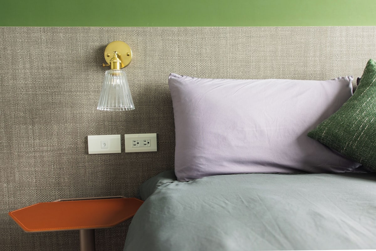 Un dormitorio ordenado con una cama, una mesita de noche, una lámpara de pared y un enchufe.