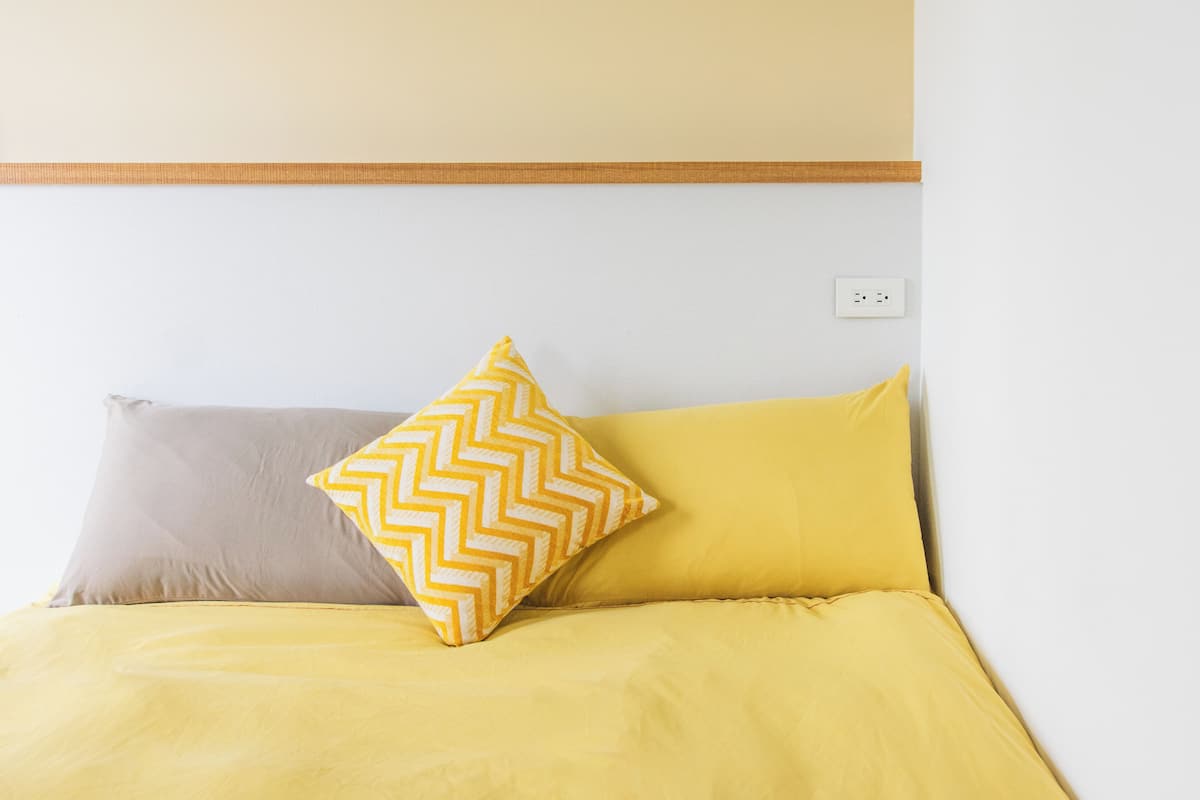 Una cama con ropa de cama amarilla y gris, un cojín estampado en patrón de rombos y un cabecero de madera.