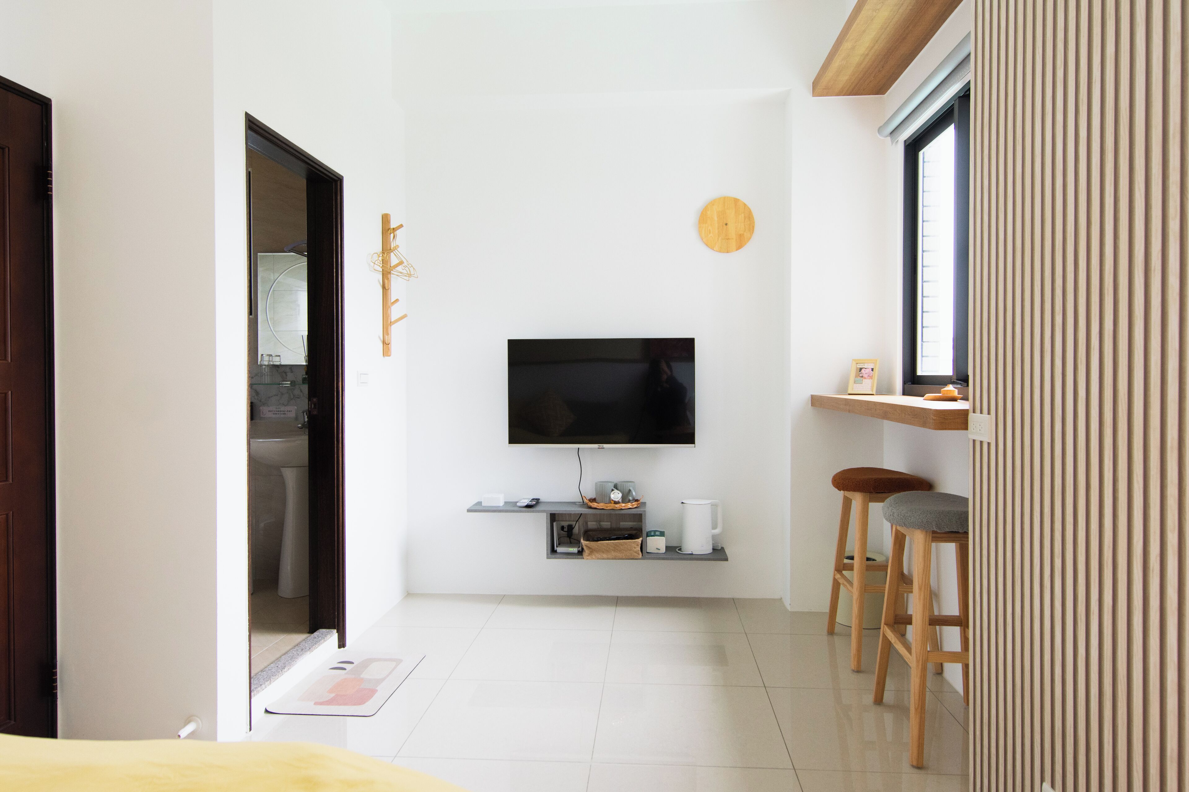 A modern living room with a flat-screen TV mounted on the wall, a small coffee table, two wooden stools, and a window with vertical blinds.