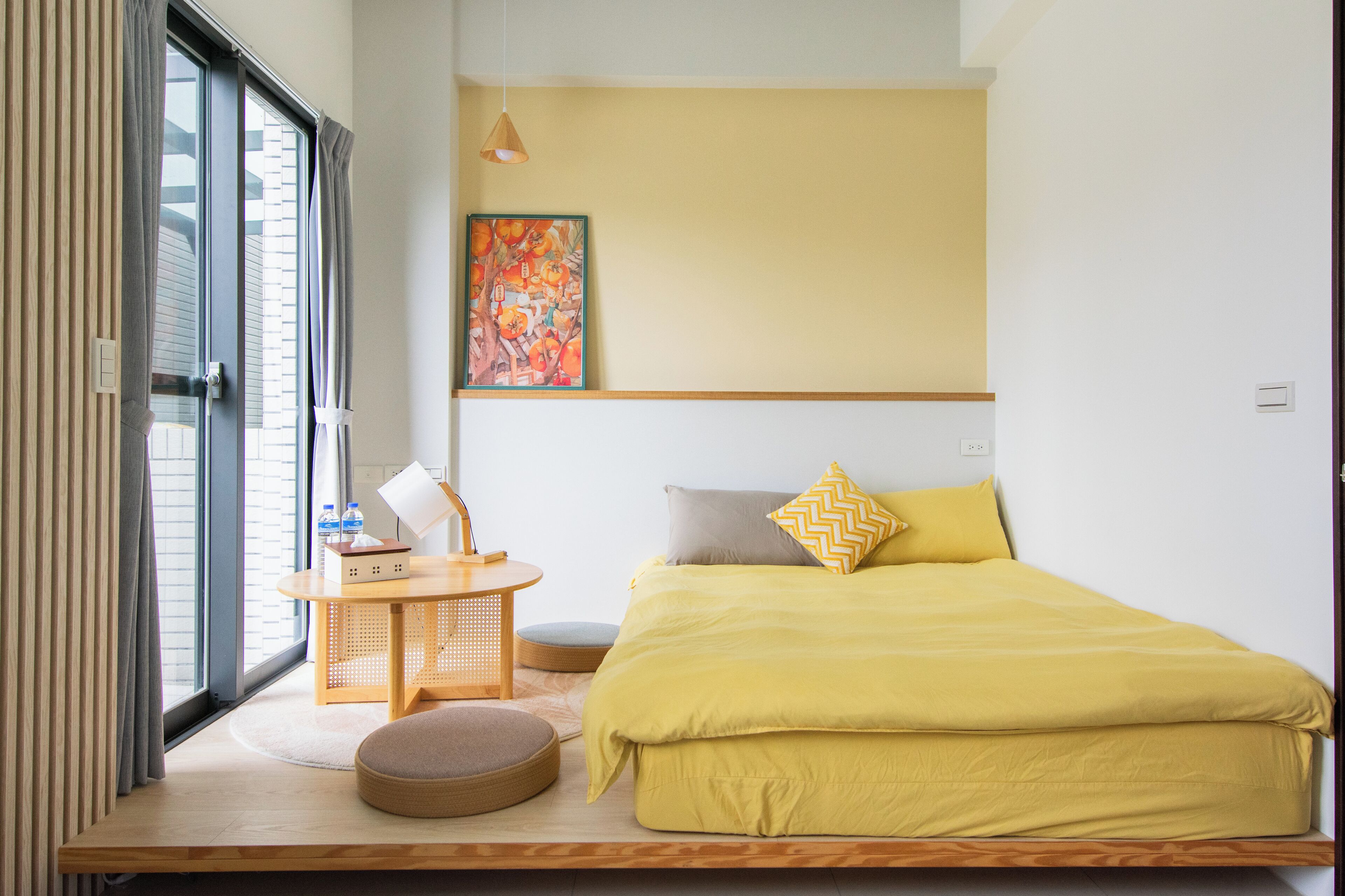 A room with a bed, a small table with a chair, a round ottoman, a painting on the wall, and a window with curtains.