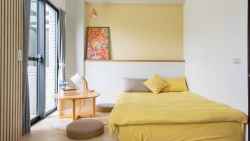 A room with a bed, a small table with a chair, a round ottoman, a painting on the wall, and a window with curtains.