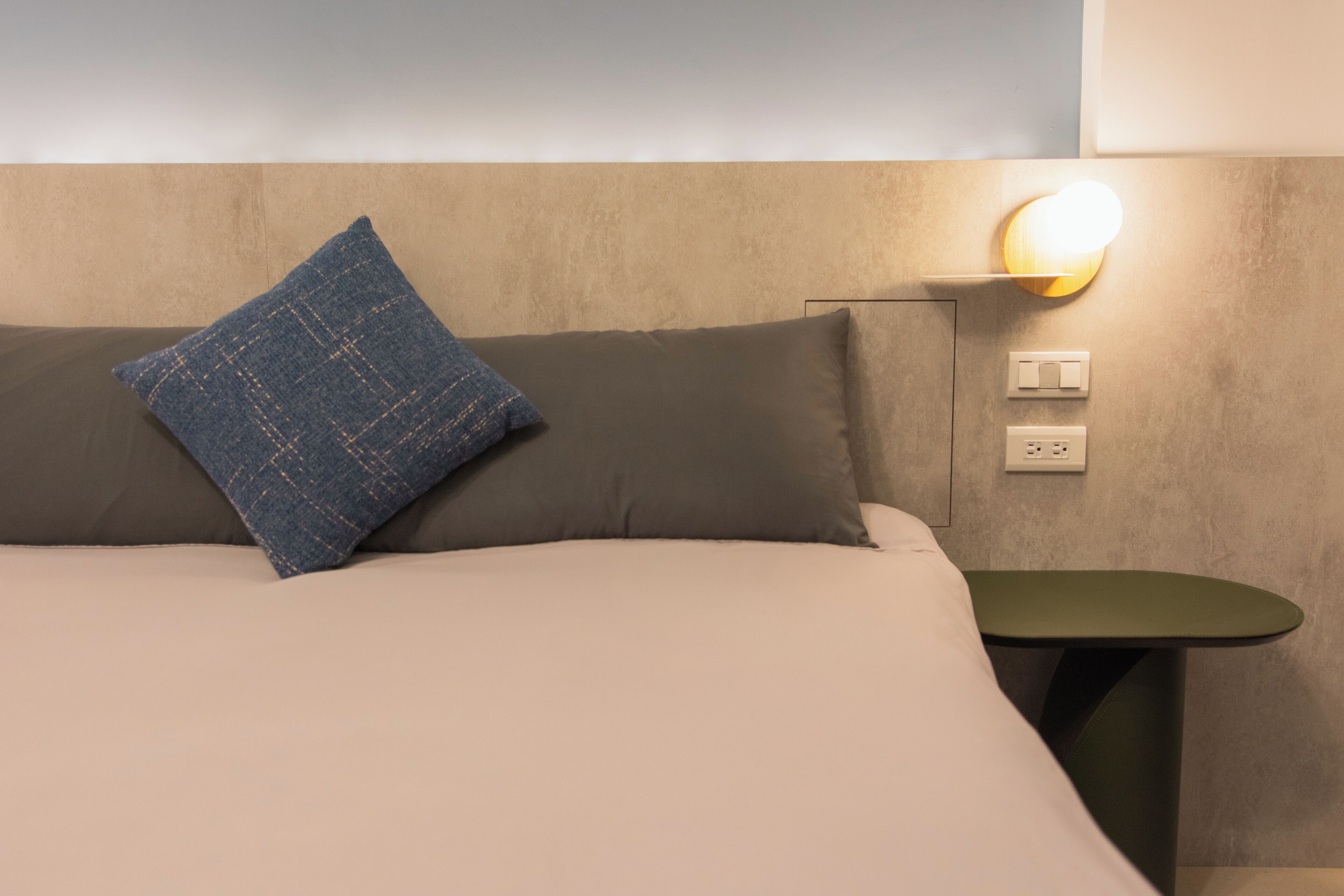 Een net opgemaakt bed met een blauw kussen, een groen nachtkastje en een wandlamp.