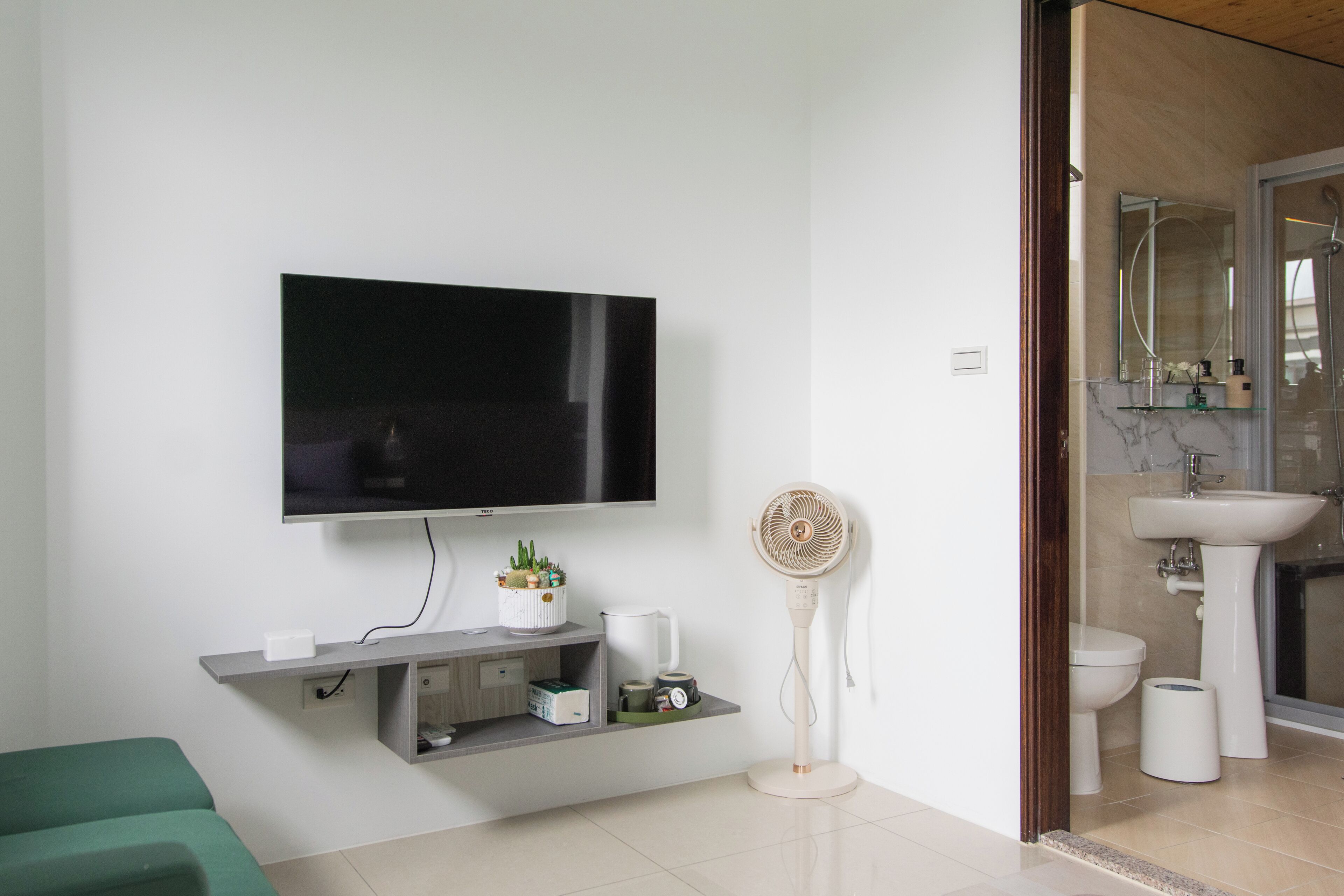 A modern living room with a flat-screen TV, a standing fan, a green couch, and a minimalist shelving unit with decorative items.