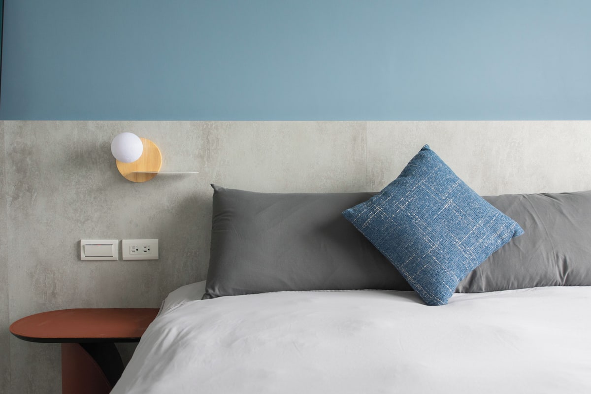 Un dormitorio con cama, almohadas grises, una almohada azul, una mesita de noche de madera y una lámpara fijada a la pared.