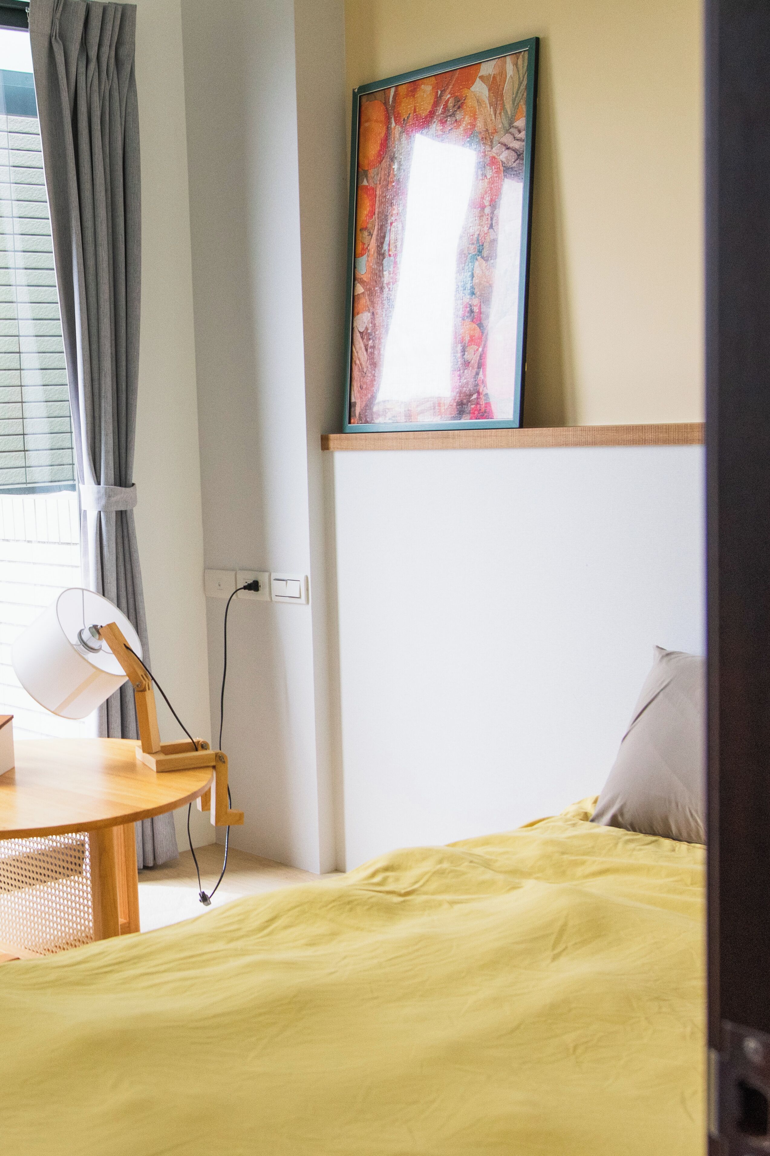 Una habitación con cama, escritorio, lámpara, un cuadro y ventana con cortinas.