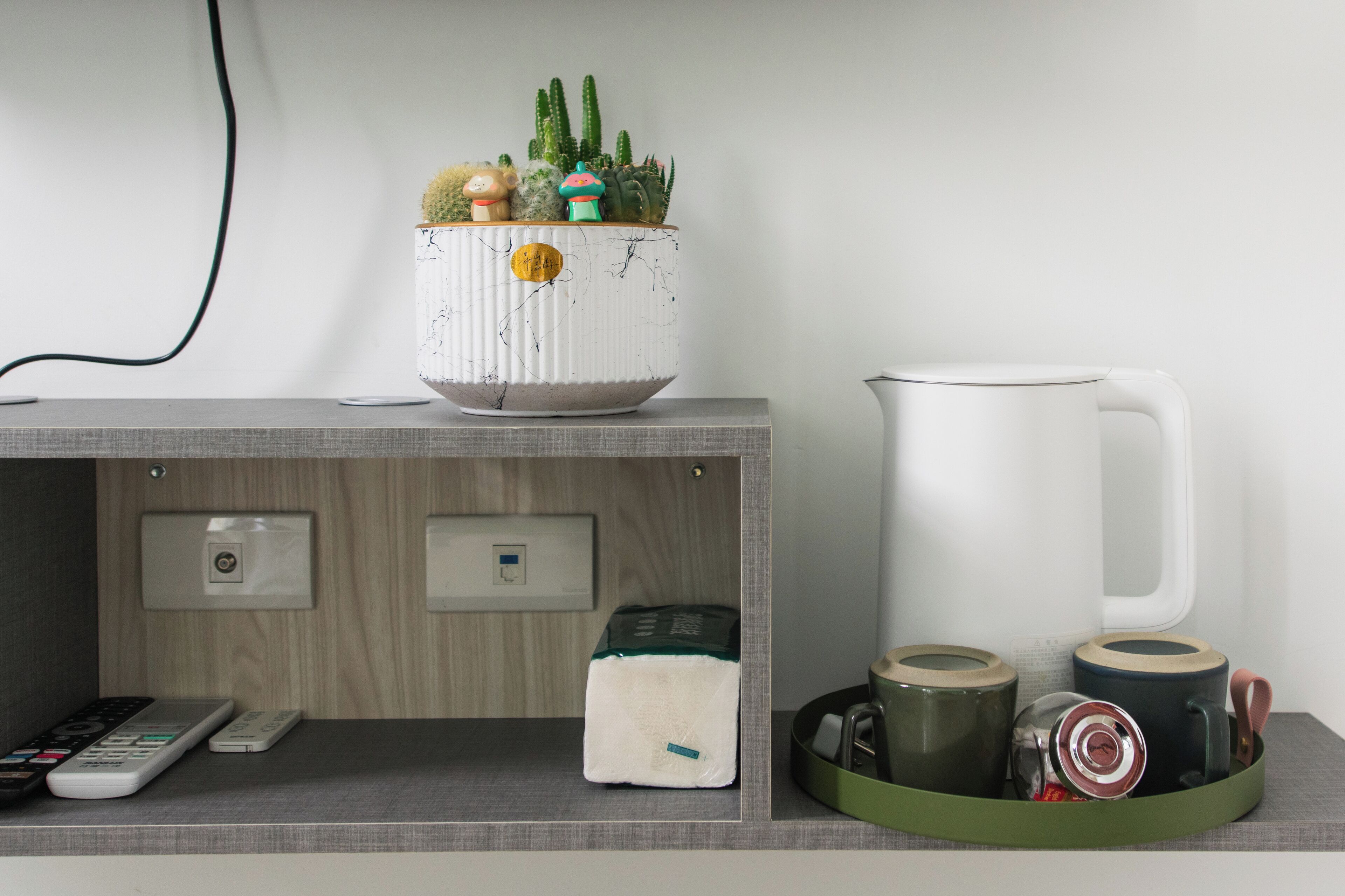 A kitchen counter with a white pot of cacti, a white pitcher, and a tray with mugs and a kettle.