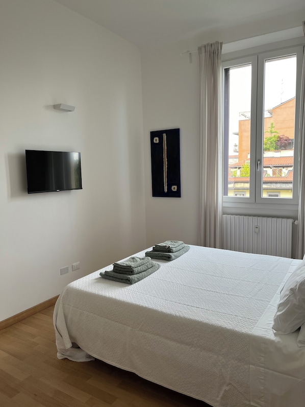 Un dormitorio con cama, televisor de pantalla plana montado en la pared, ventana con cortinas y radiador.
