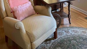 Quarto com cama de madeira, uma cadeira com almofada rosa, mesa e tapete estampado.