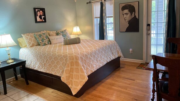 Un dormitorio con cama, dos mesitas de noche, una silla, una araña y una ventana con cortinas.