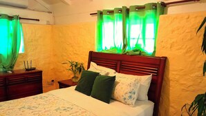 Un lit bien fait, recouvert d’une couvre-lit à motifs floraux, avec une tête de lit en bois et des oreillers verts et blancs. On y trouve une table de chevet ornée d’une plante et d’une petite lampe de table.