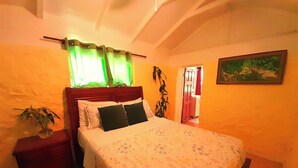 Une chambre à coucher avec un lit, une table de chevet, une plante en pot, un tableau accroché au mur et une porte donnant sur une autre pièce.