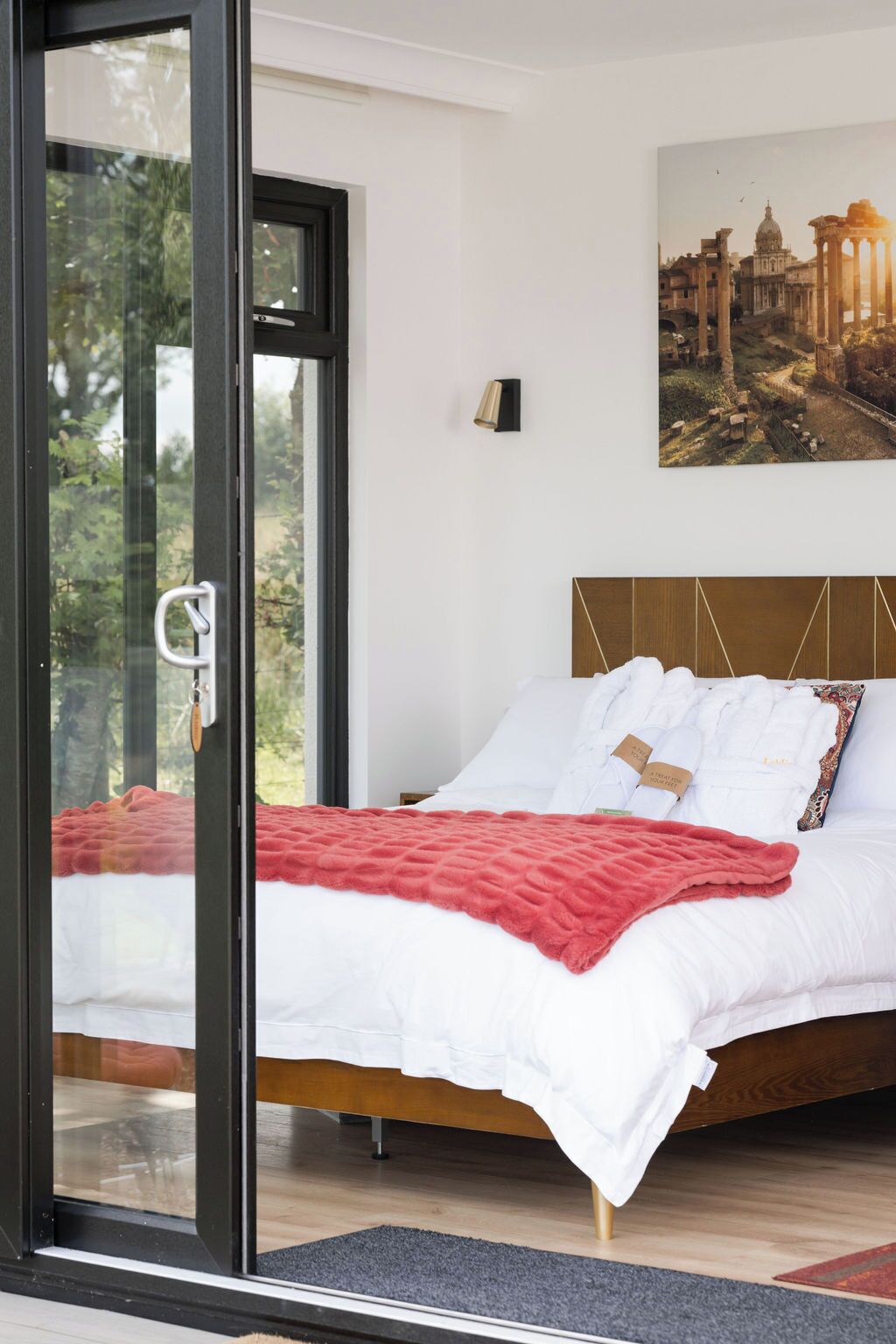 A modern bedroom with a glass door leading to a balcony, a large bed with white bedding, a red throw blanket, and a cityscape painting on the wall.