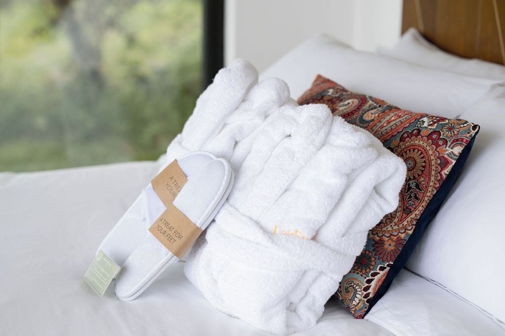A neatly made bed with white bedding, a colorful patterned pillow, and a pair of white bathrobes with a tag.