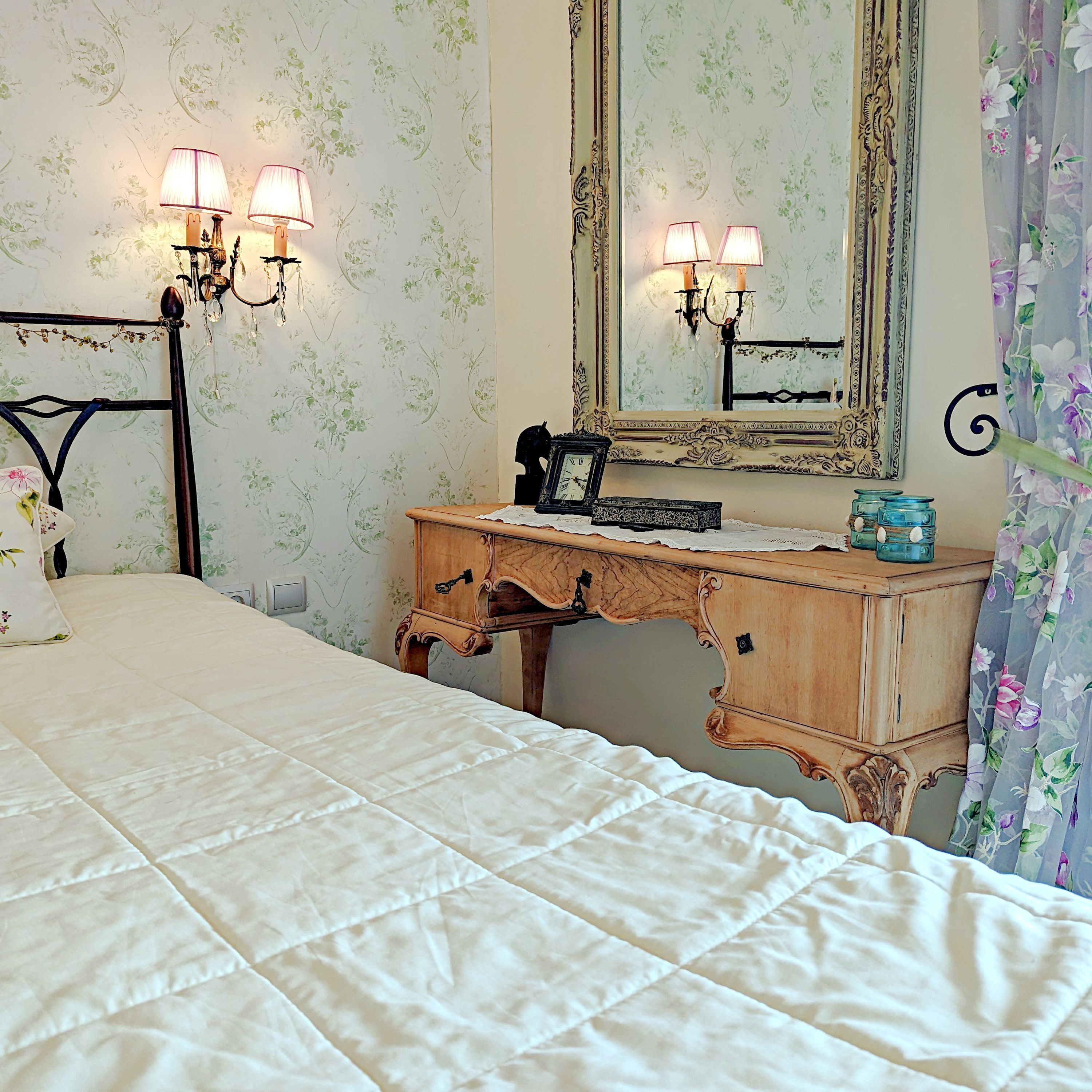 A bedroom with a bed, a wooden dresser, a mirror, and wall sconces.