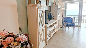 Ein Wohnzimmer mit Holzboden, einer weißen Unterhaltungsanlage, einem blauen, gemusterten Sessel und Meerblick durch ein großes Fenster.