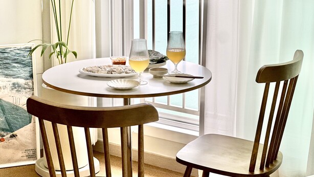Una mesa redonda con mantel blanco, dos sillas de madera, un vaso de bebida, un plato de comida y un cuenco pequeño.