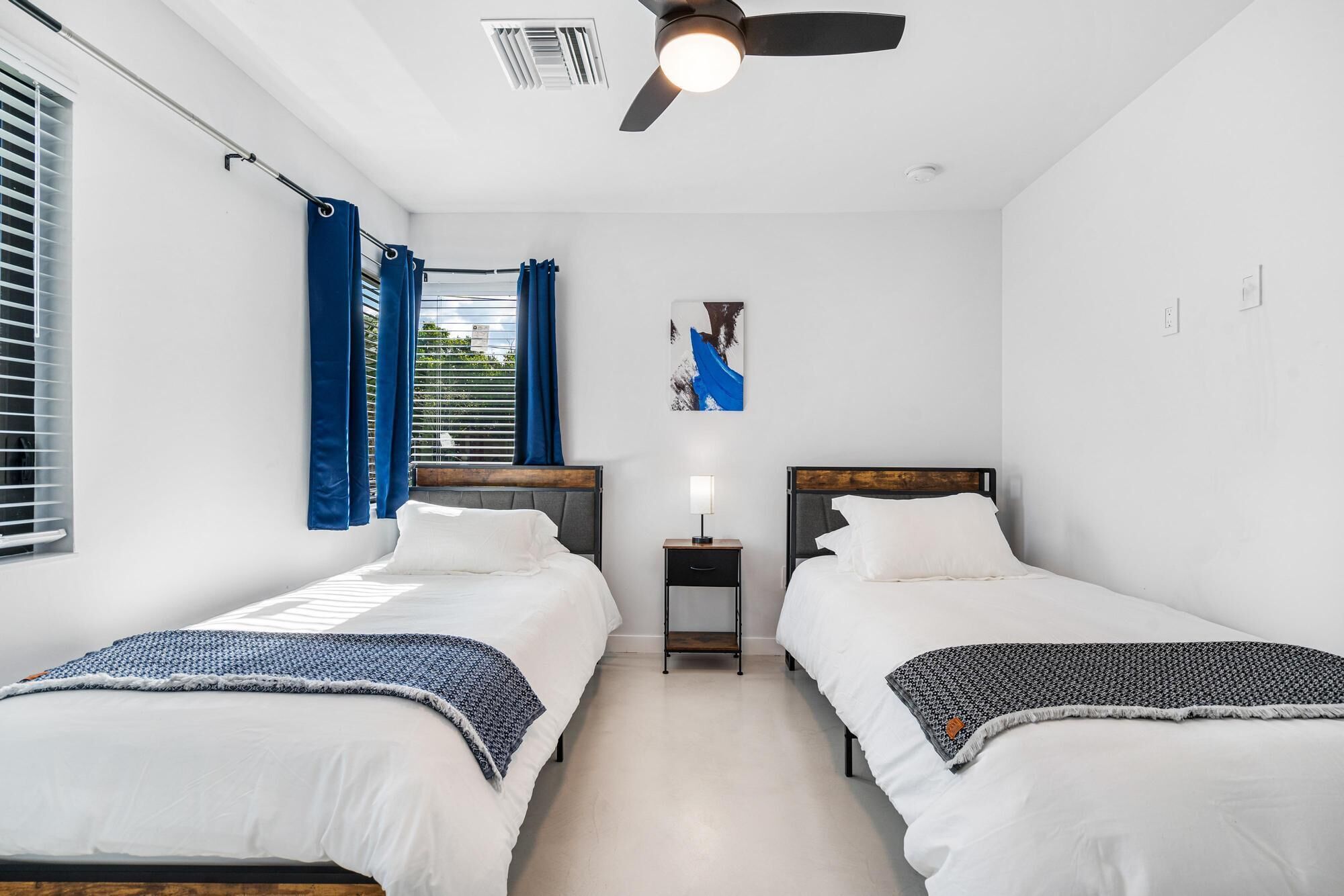 A hotel room with two single beds, a ceiling fan, a nightstand with a lamp, and a window with blinds.