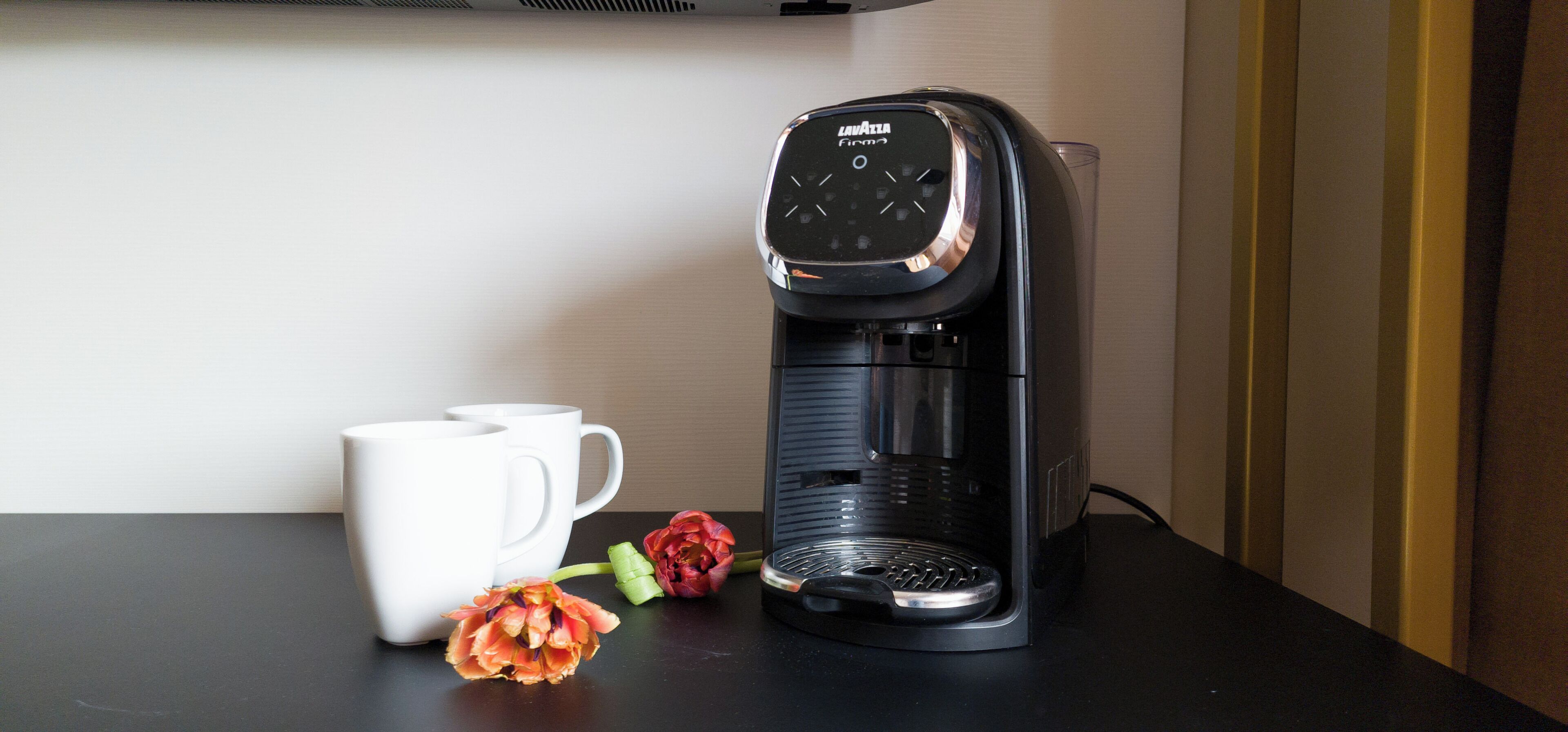 A black coffee maker with a digital display, two white cups, and a red flower on a dark surface.