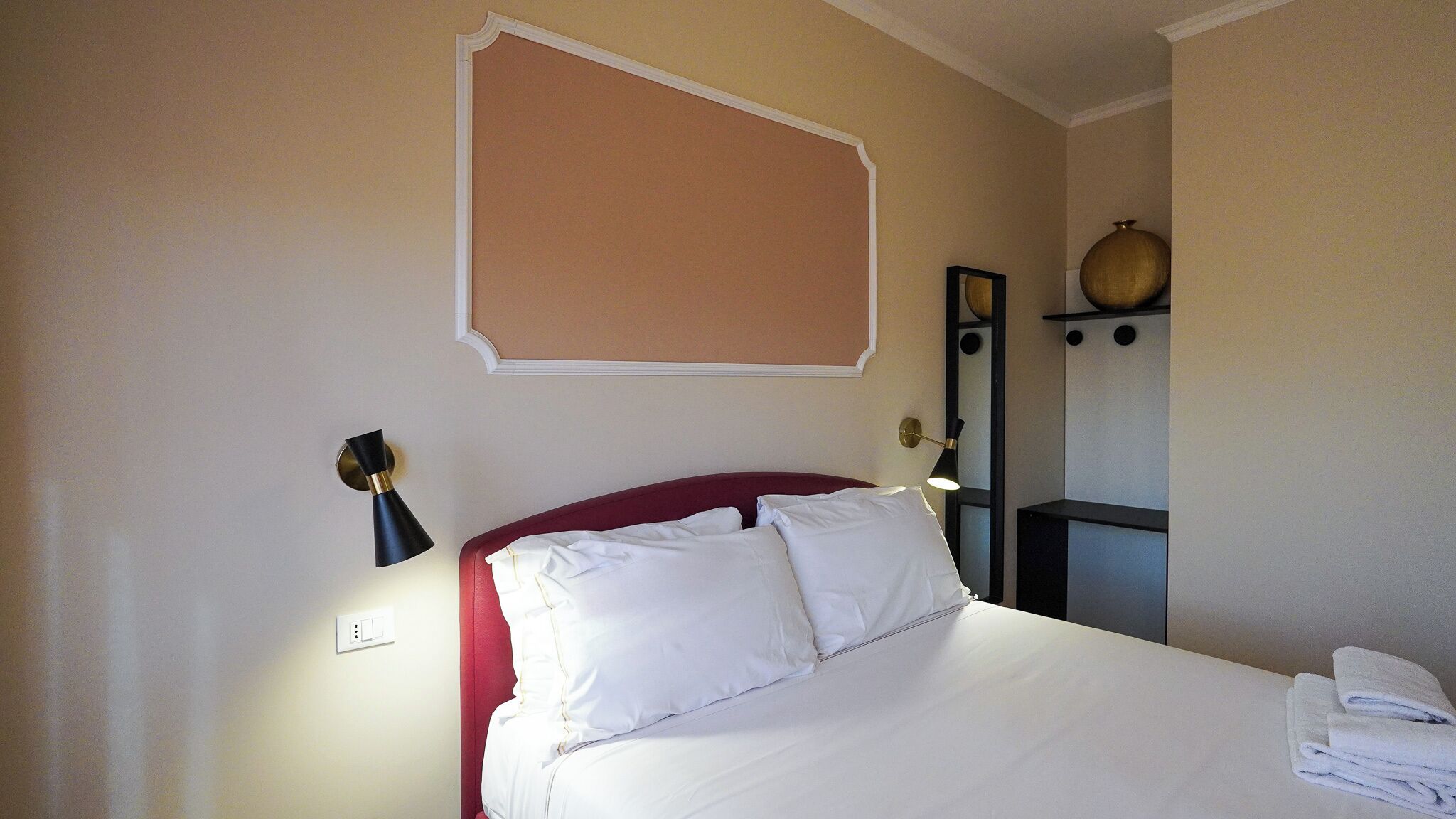 Ein Hotelzimmer mit einem Bett, einer Nachttischlampe, einem Wandregal mit Dekoration und einer Steckdose.