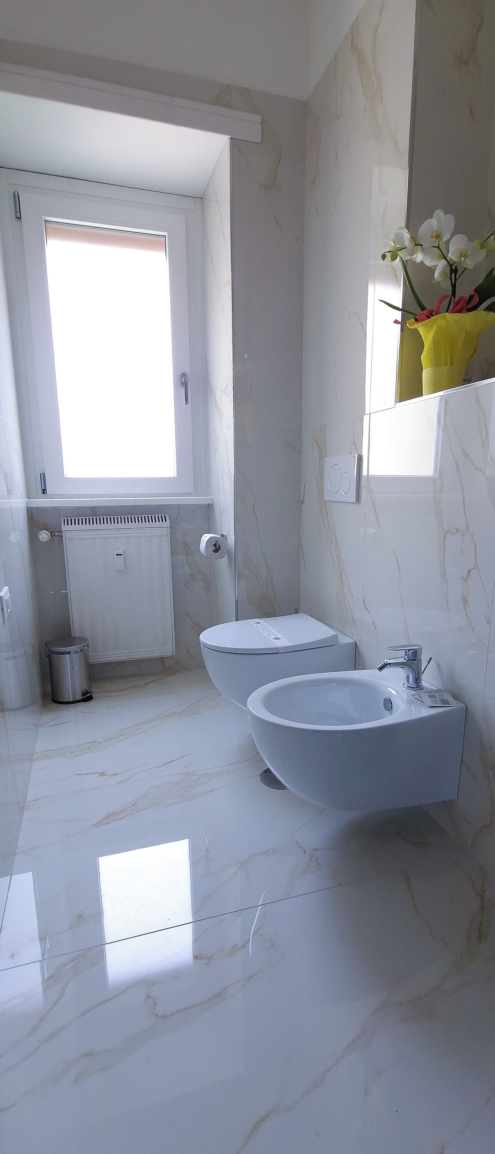 A modern bathroom with two sinks, a toilet, a window, and a mirror with a vase of flowers.