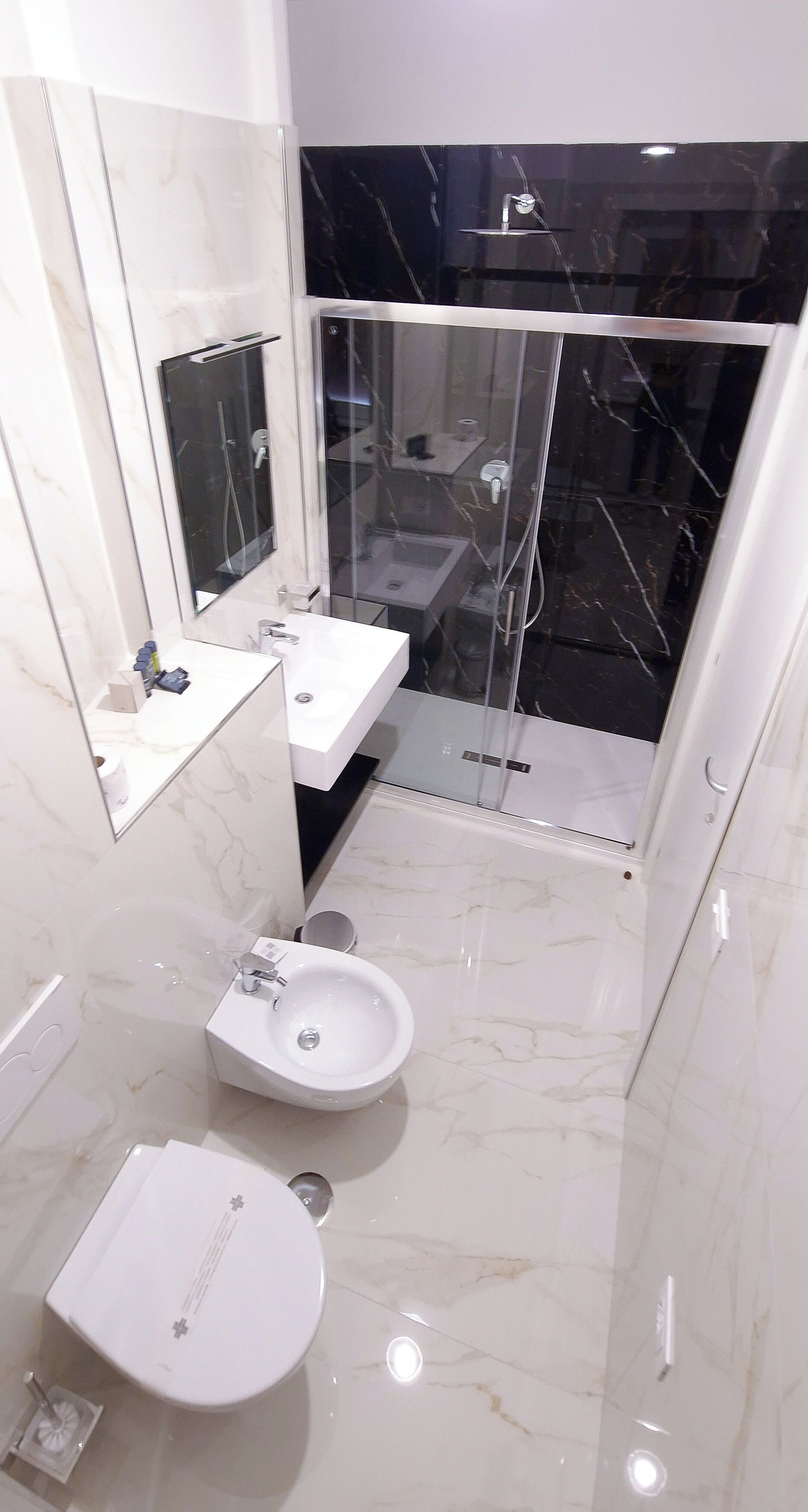 A modern bathroom with a white marble floor, a large mirror, a white sink, a bidet, and a glass-enclosed shower area.