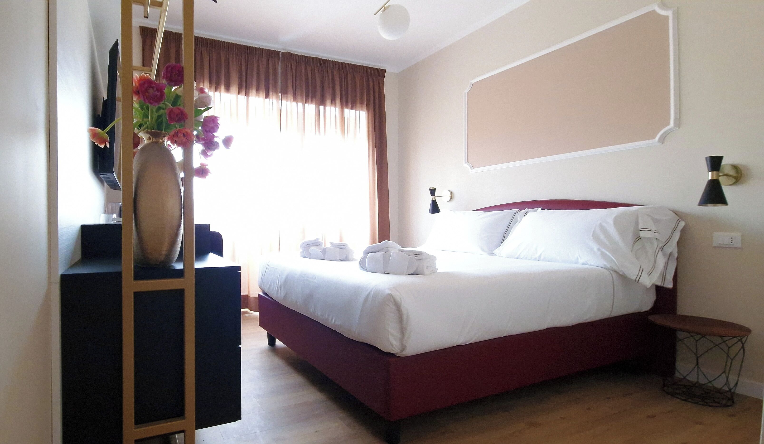 A hotel room with a bed, a desk, a vase with flowers, and a window with curtains.