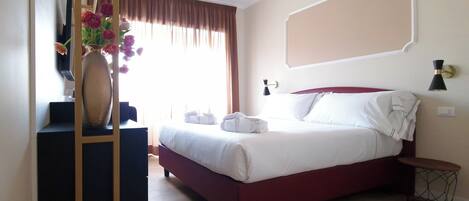 A hotel room with a bed, a desk, a vase with flowers, and a window with curtains.