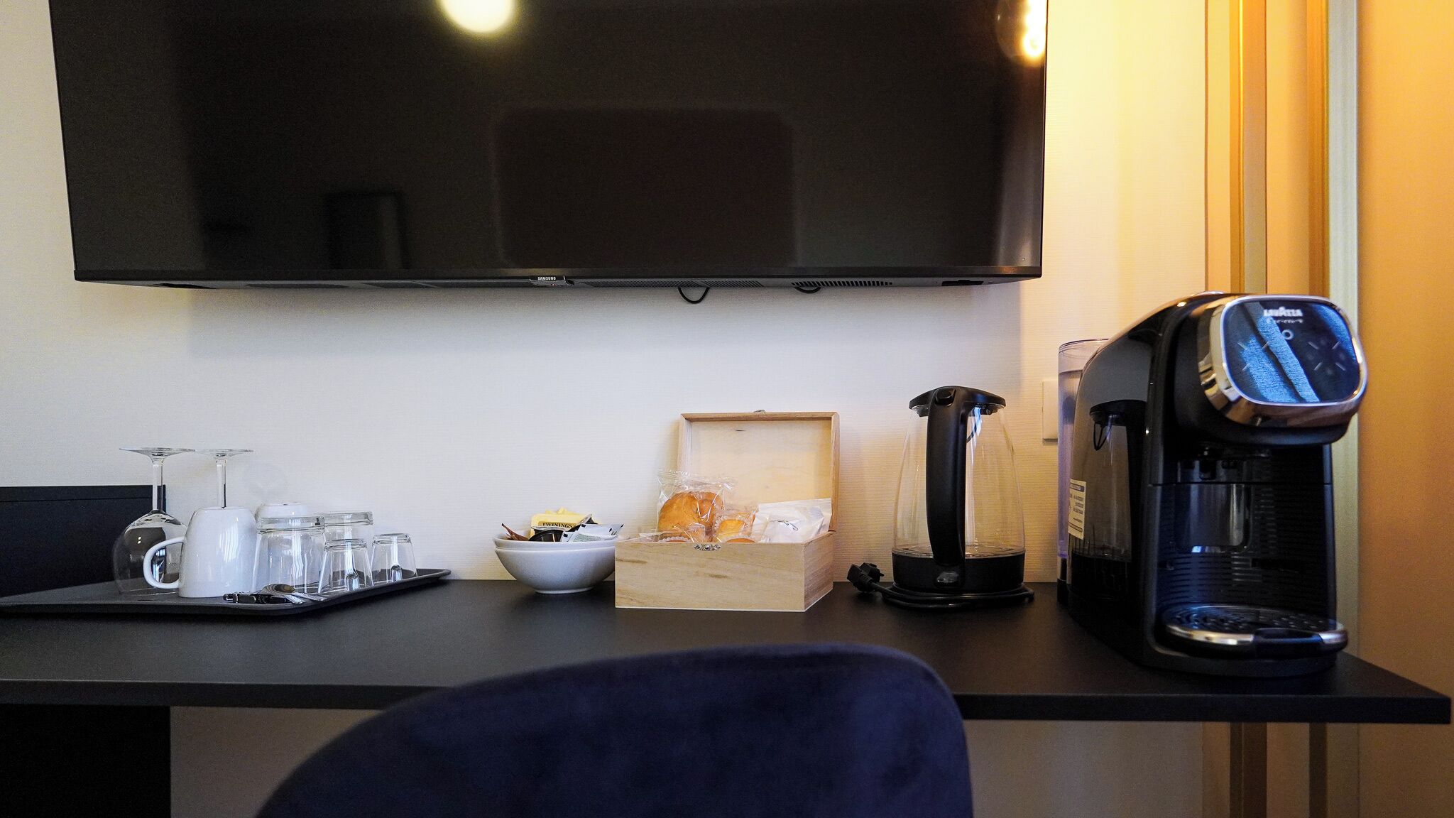 A hotel room with a flat-screen TV mounted on the wall, a coffee maker, a glass carafe, and a tray with glasses and a bowl of bread.